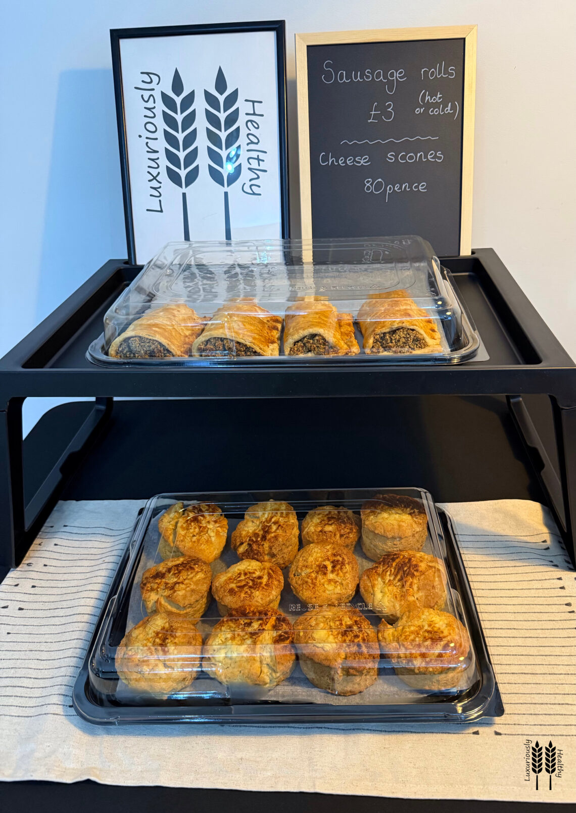 Sake display of sausage rolls and cheese scones. Two tiers, a board with a logo and a board with prices